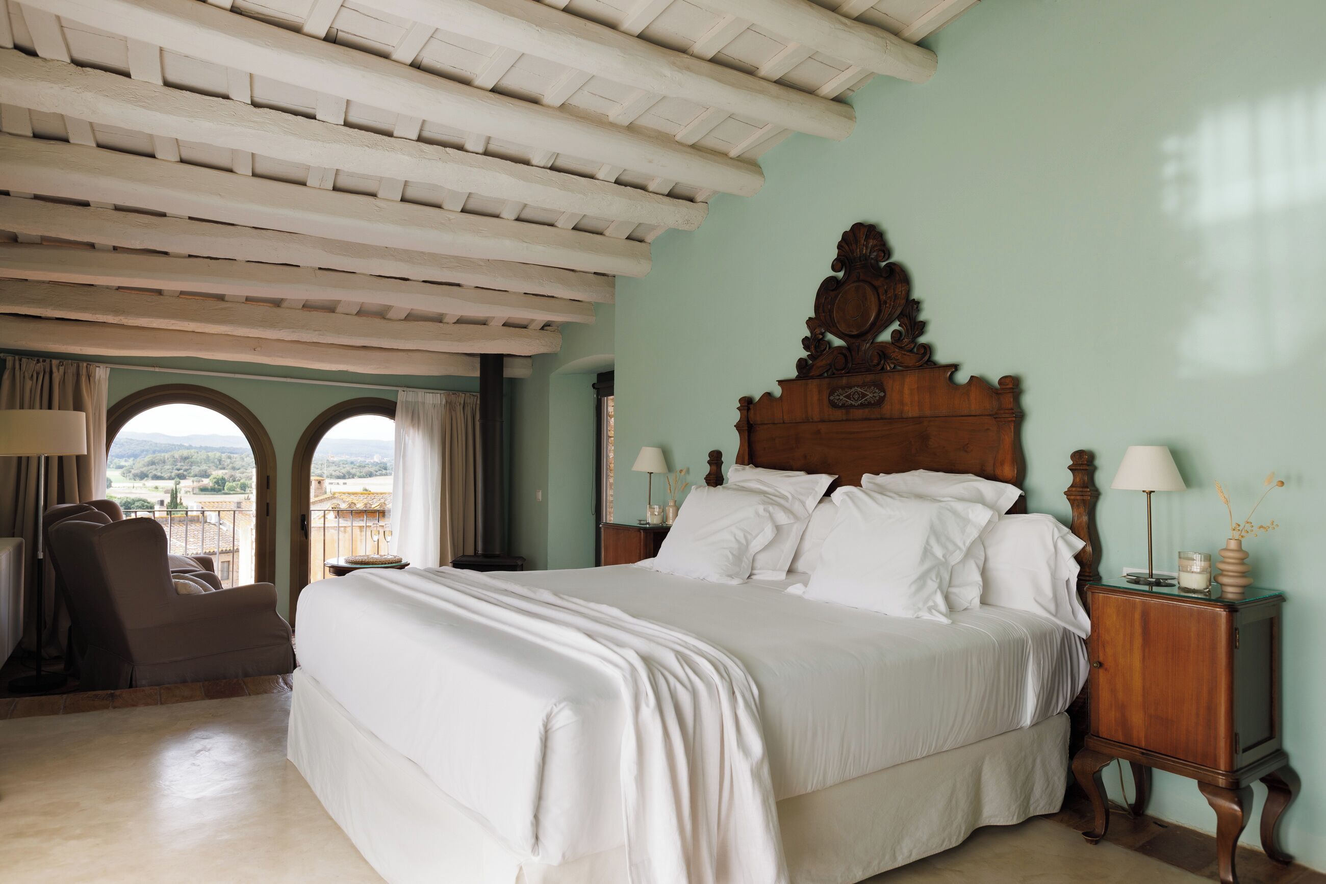 Un dormitorio con una cama grande, una mesita de noche de madera, una silla y vistas a través de ventanas de arco.