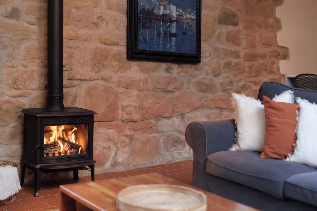 A stone wall, a black wood-burning stove, a framed picture, a gray sofa, and a wooden coffee table.