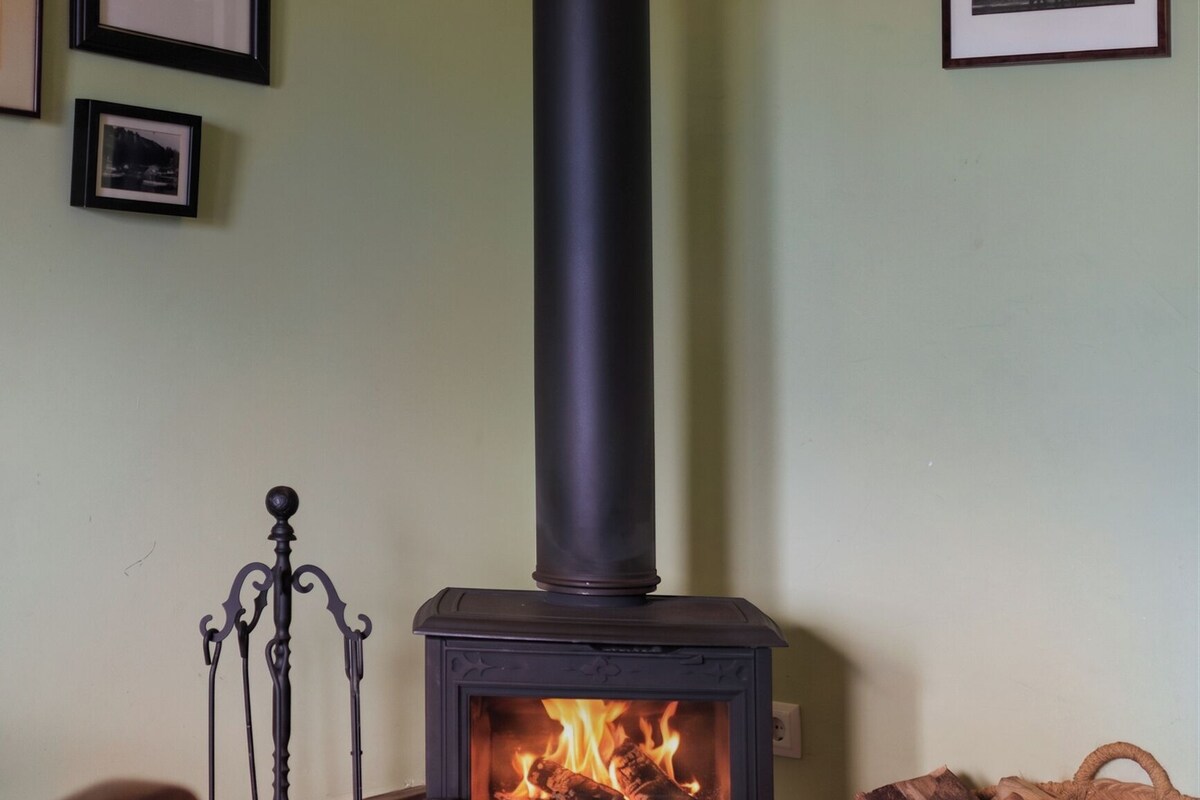 A room with a wood-burning stove, a chair, a table with a cup, and framed pictures on the wall.