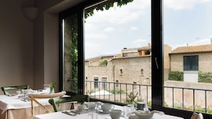 A dining area with tables set for two, a view of buildings through large windows, and a balcony railing. - Encís d Empordà (Corca)