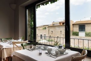 A dining area with tables set for two, a view of buildings through large windows, and a balcony railing.
