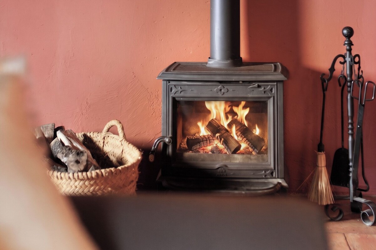 A wood-burning stove with a chimney, a basket of firewood, and a broom.