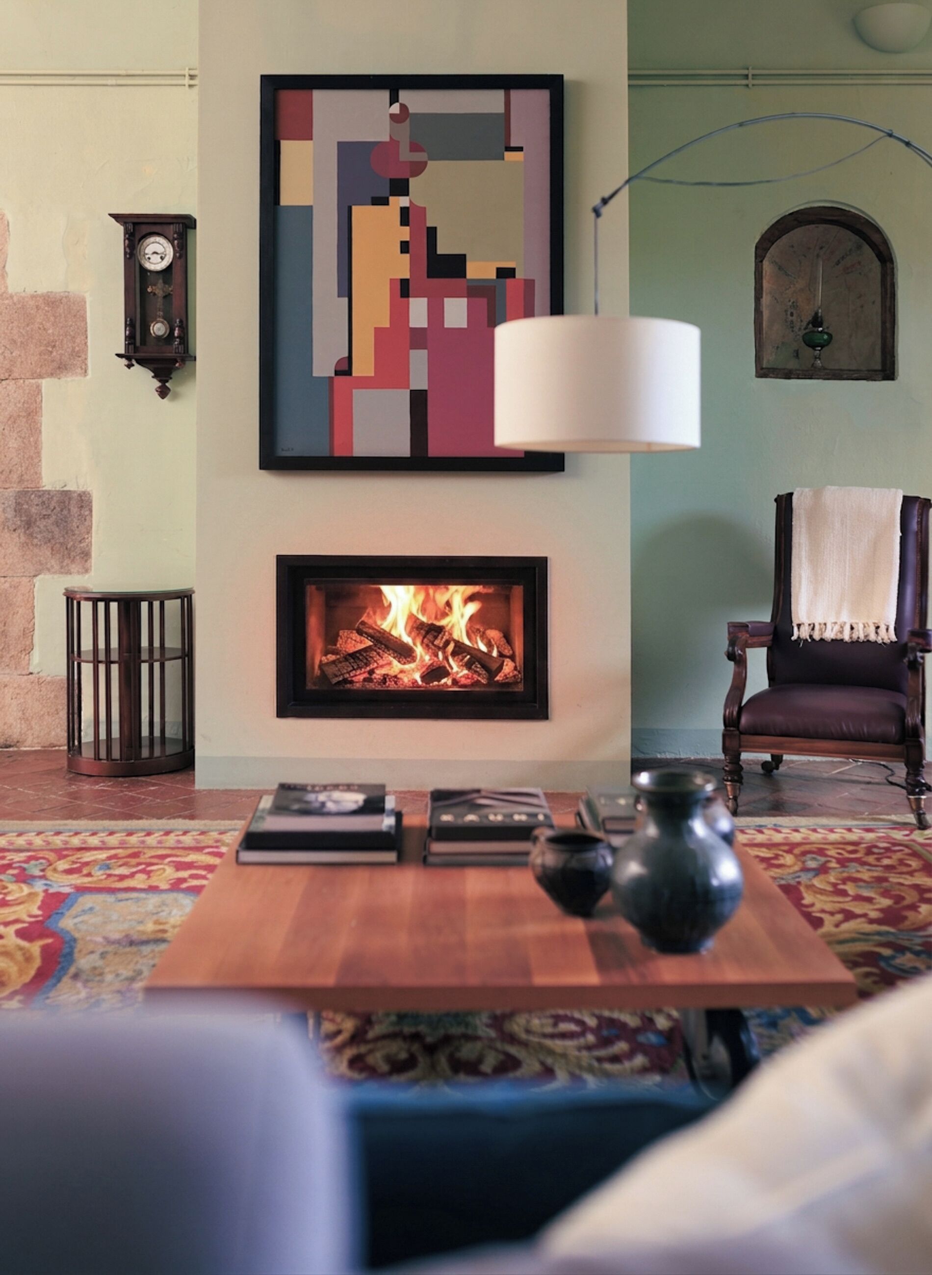 A living room with a fireplace, a clock, a chair, and a table with books and a vase.