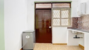 A compact kitchen with a brown door, a small refrigerator, a window with a mesh screen, and a tiled backsplash. - Hotel Vivenda Rebelo (Panaji)
