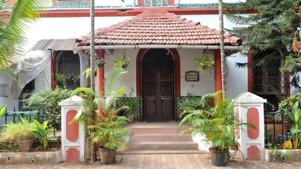 A building with a red-tiled roof, a central entrance with a dark door, and potted plants around the entrance. - Hotel Vivenda Rebelo (Panaji)