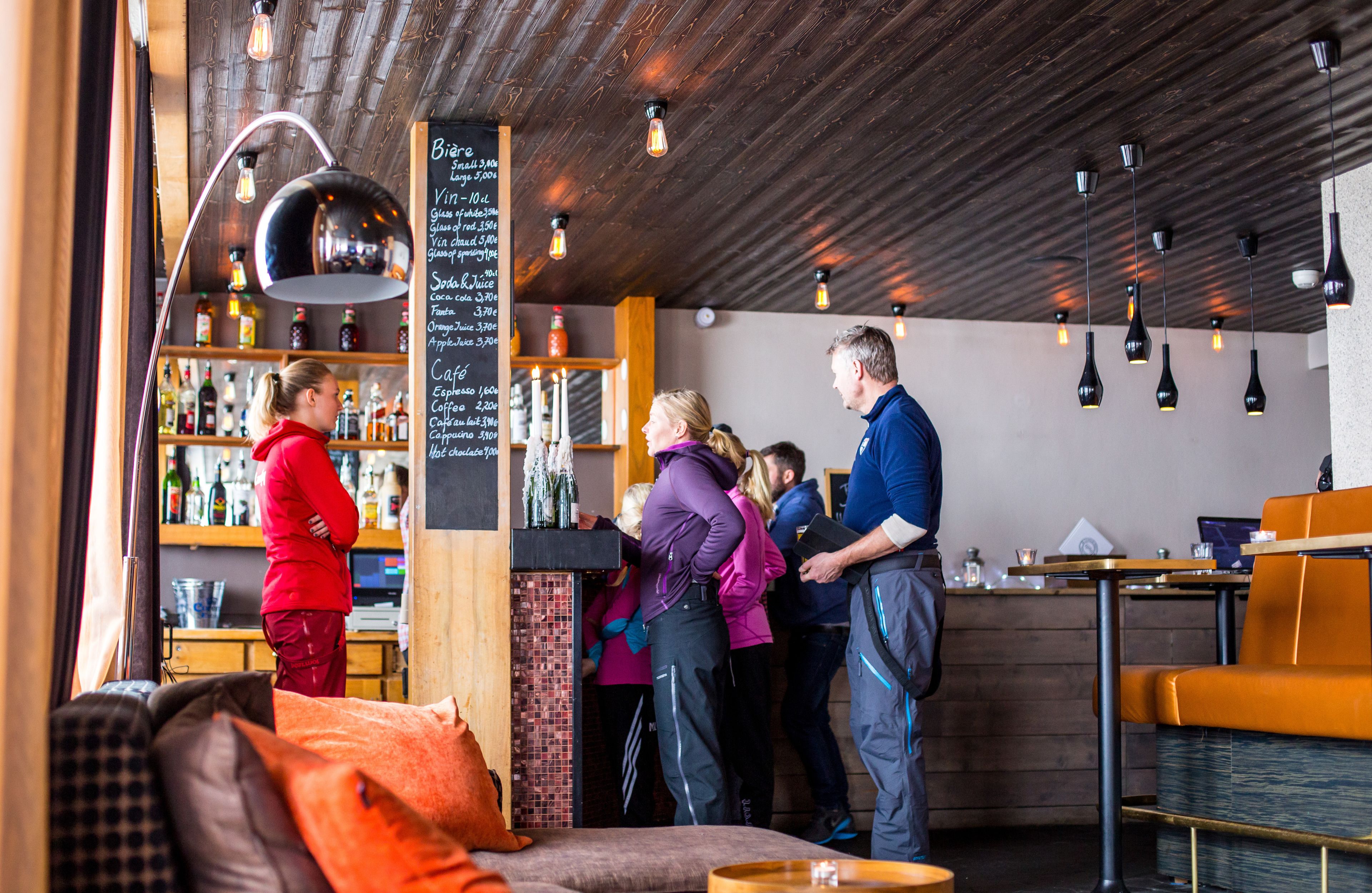 A bar area with a wooden ceiling, hanging pendant lights, and a chalkboard menu listing drinks.