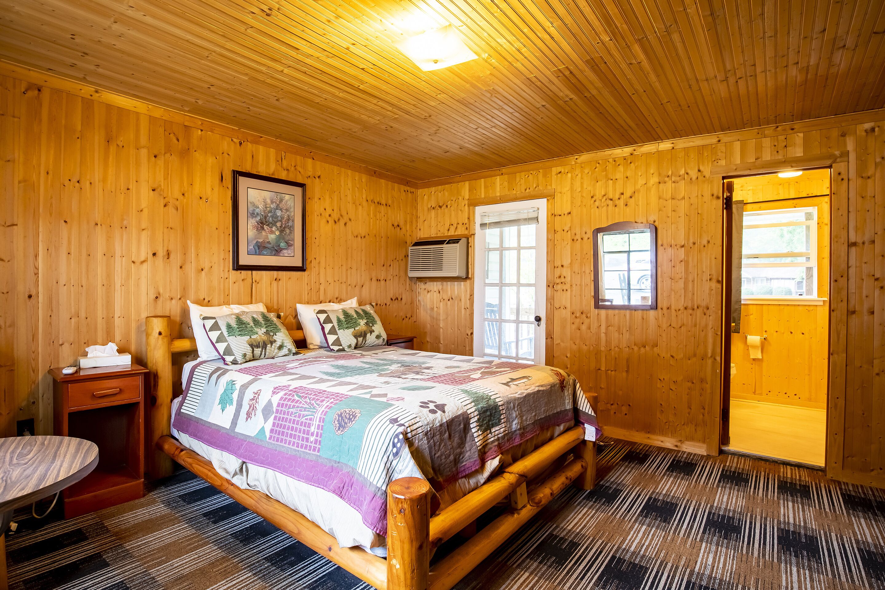 A wooden-paneled bedroom with a bed, bedside table, and a door leading to another room.