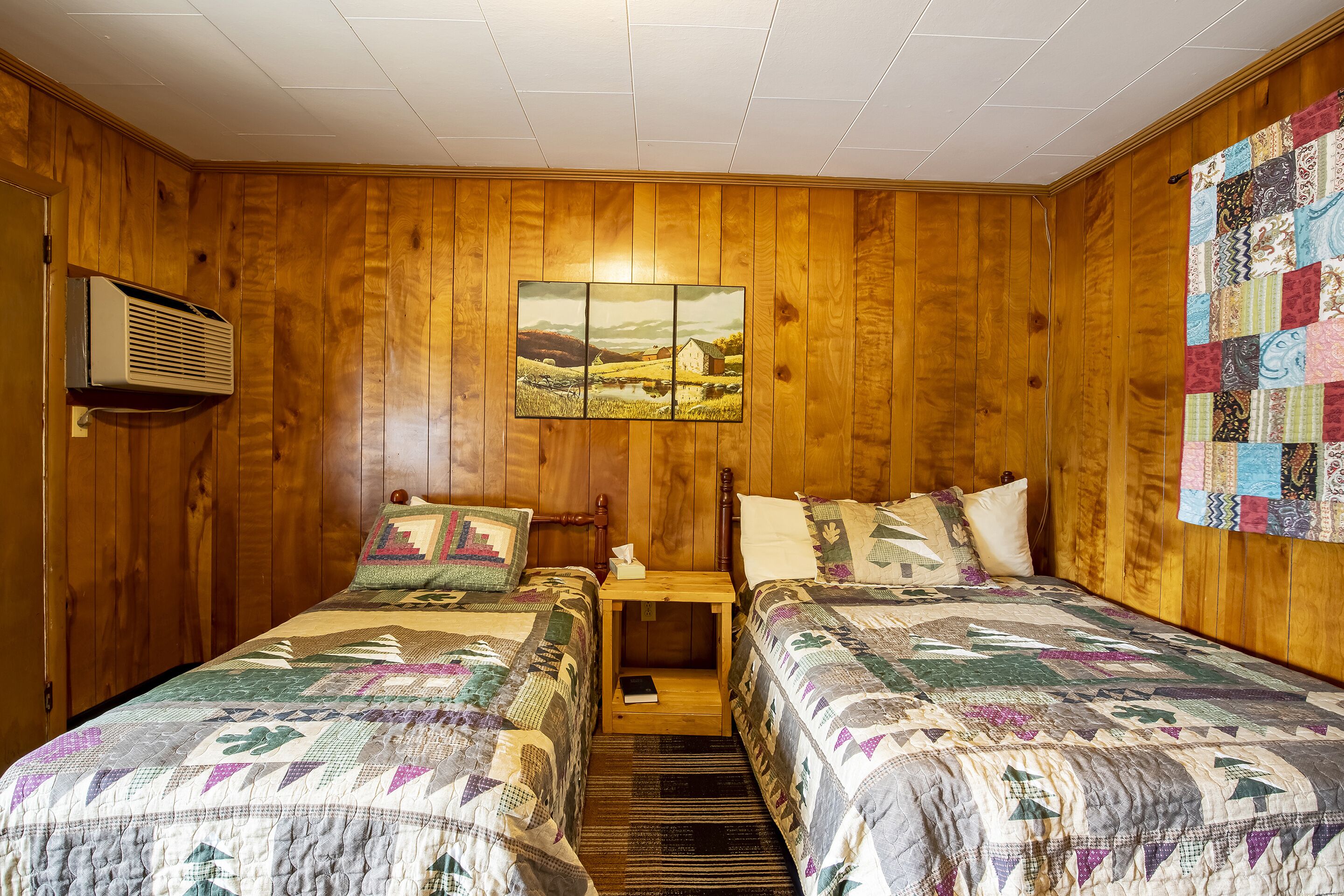 A room with wooden walls, two beds with quilts, a nightstand with a lamp, a quilted wall hanging, and a picture of a landscape.