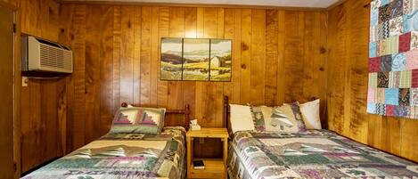 A room with wooden walls, two beds with quilts, a nightstand with a lamp, a quilted wall hanging, and a picture of a landscape.