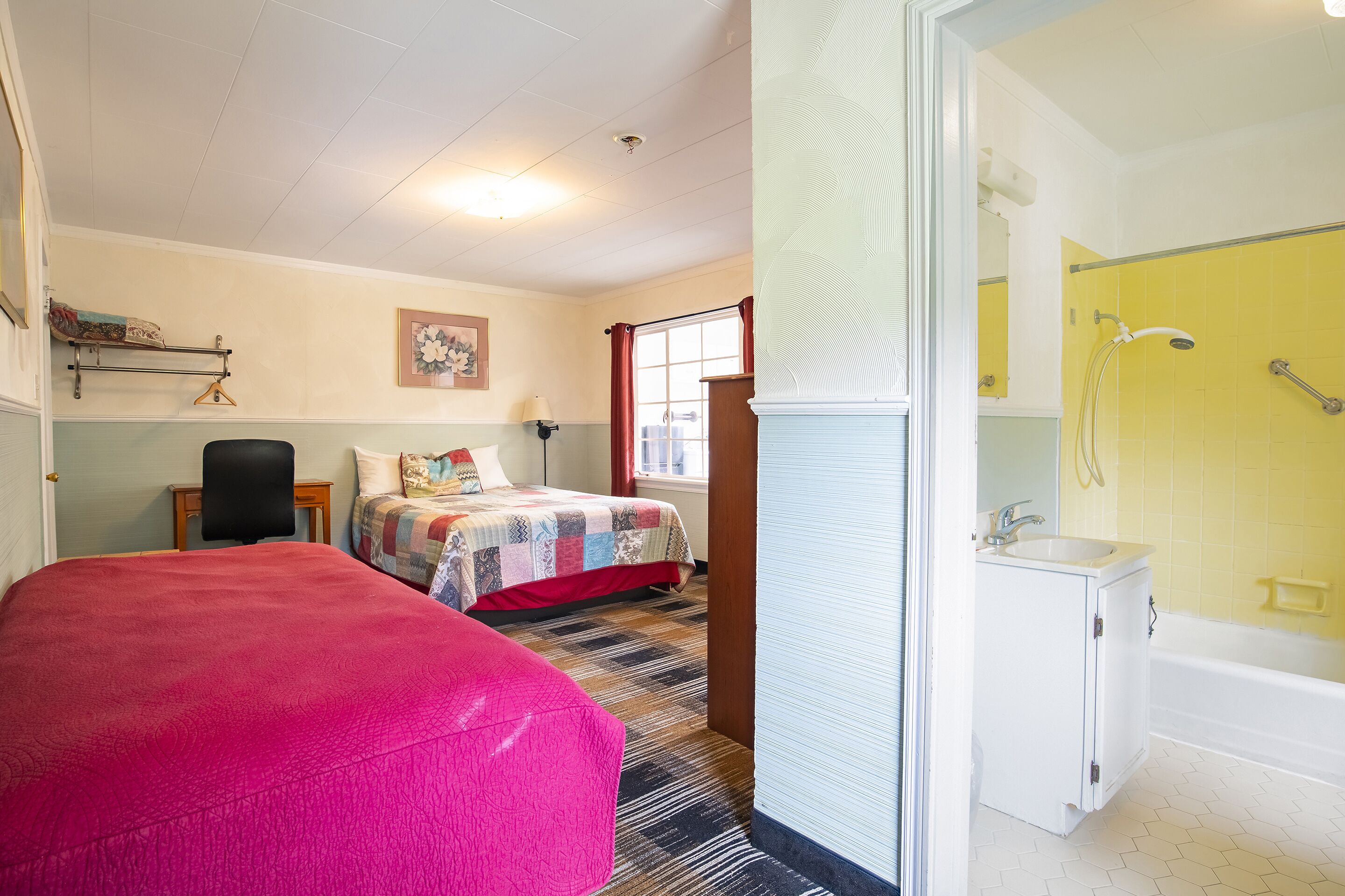 A room with a bed, a chair, a small table, a window with red curtains, and a bathroom with a yellow-tiled shower and white sink.