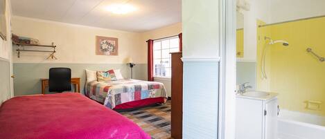 A room with a bed, a chair, a small table, a window with red curtains, and a bathroom with a yellow-tiled shower and white sink.