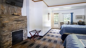 A hotel room with a stone fireplace, a bed with a blue quilt, a television mounted on the wall, and a window with curtains.