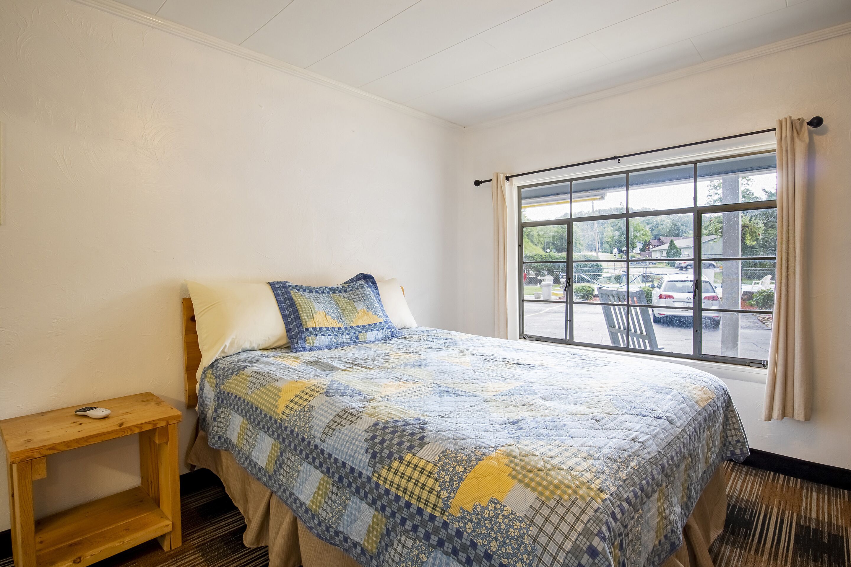 A bedroom with a bed, a wooden nightstand, and a window with a view of a parking lot.