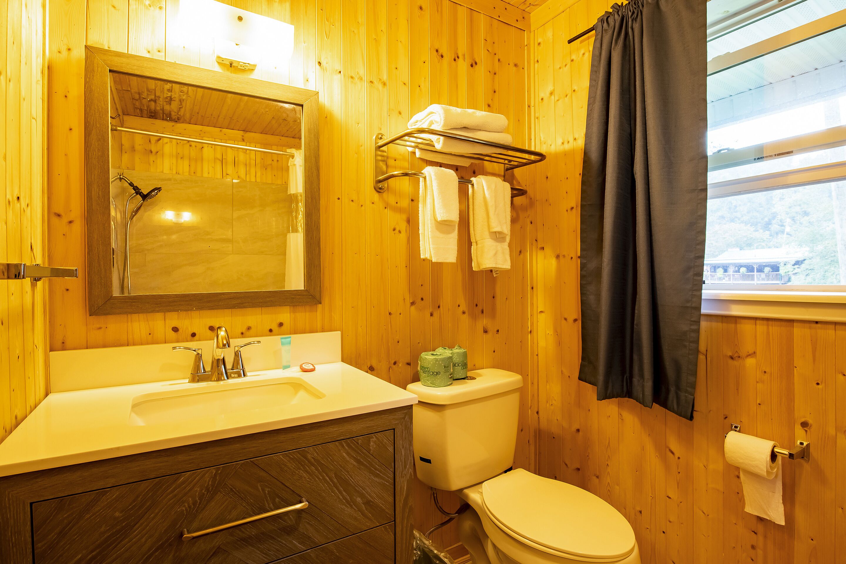 Une salle de bain rustique, aménagée dans une cabane en bois, comprenant un lavabo, un miroir, un porte-serviettes, des toilettes et une fenêtre donnant sur un paysage agréable.
