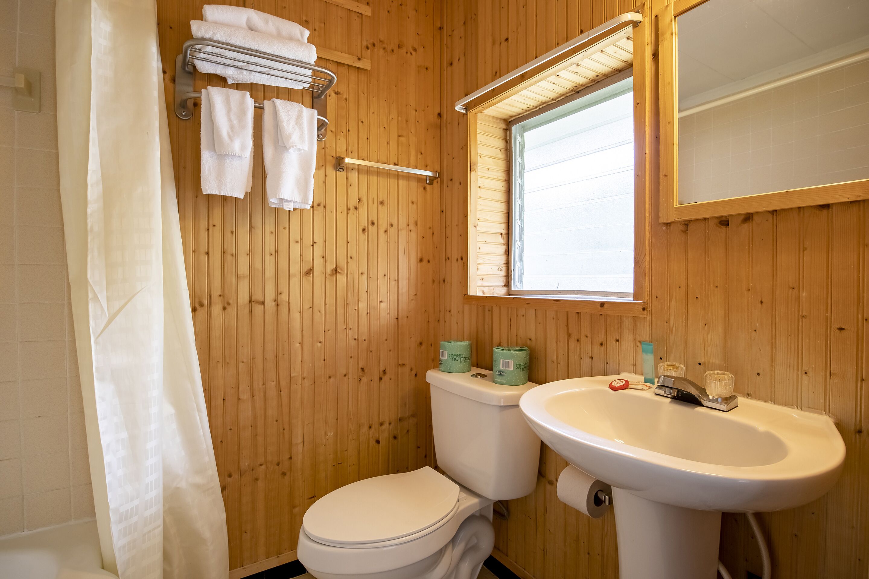 A wooden-paneled bathroom with a white toilet, a sink, and a window above the mirror.
