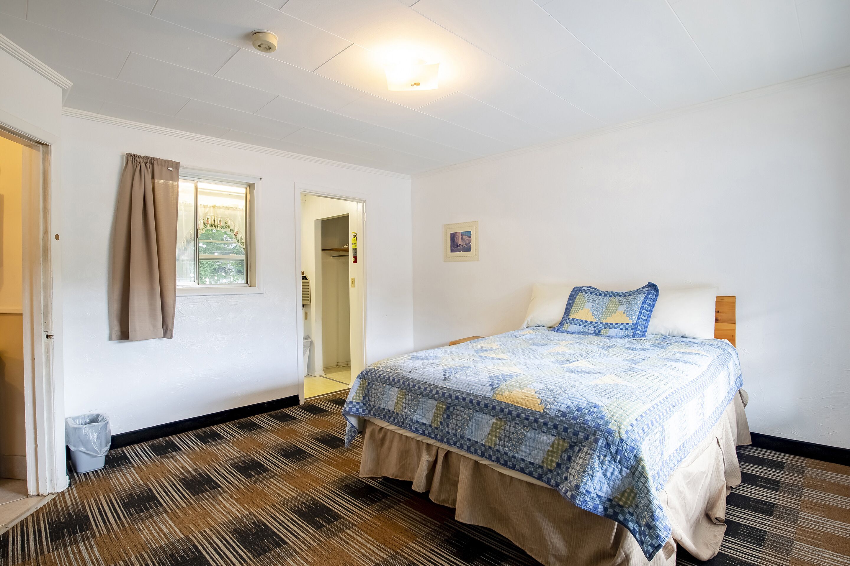 A bedroom with a bed, a window with curtains, a door, and a patterned carpet.