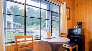 A wooden interior with a round table and two chairs, a window with a view of a parking lot and trees, and a small black appliance on the wall.