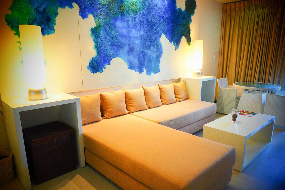 Quarto de hotel moderno com um sofá grande, uma mesa pequena e uma mesa de centro de vidro. Há uma luminária em cada lado do sofá e um grande quadro abstrato na parede.