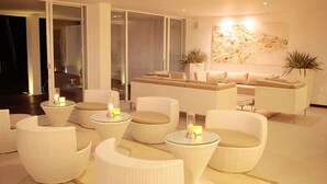 Una sala de estar moderna con muebles de mimbre blanco, incluyendo sofás y sillones, y una mesa de centro de vidrio. Hay velas sobre las mesas, y un cuadro abstracto grande en la pared.