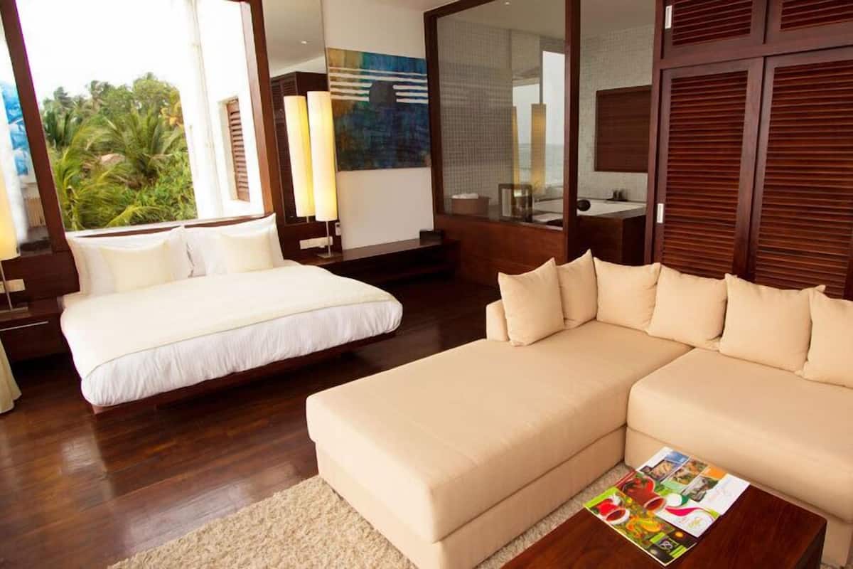 Quarto de hotel moderno com cama, sofá, piso de madeira, espelho e janela com vista para o verde.