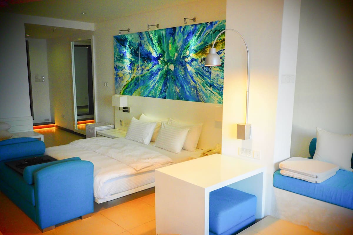 Quarto de hotel moderno com uma cama grande, uma escrivaninha, um sofá azul e um quadro abstrato vibrante na parede.