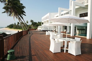 A wooden deck with white wicker furniture and umbrellas, overlooking a beach with palm trees and a modern building with balconies. - Michelle Boutique Hotel (Balapitiya)