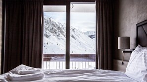 Quarto de hotel com uma cama grande, roupa de cama branca, vista de uma paisagem montanhosa coberta de neve através de uma varanda e uma luminária na parede.