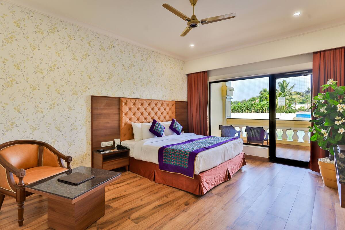 Une chambre d’hôtel avec un grand lit, une tête de lit en bois, une table de chevet, un bureau, une chaise, un ventilateur de plafond et un balcon avec vue sur les palmiers.