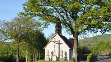 Une petite église surmontée d’une croix, entourée d’arbres et d’un mur de pierre.
