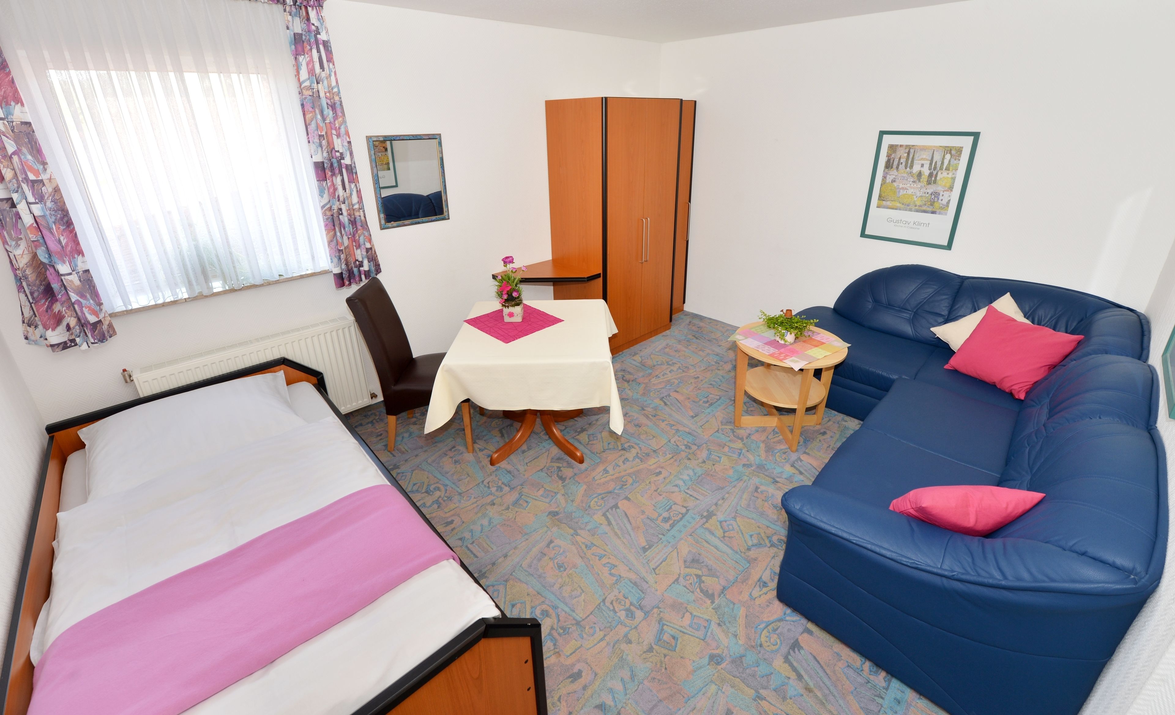 A room with a bed, a blue sofa, a small table with a plant, a chair, a wardrobe, and a window with curtains.