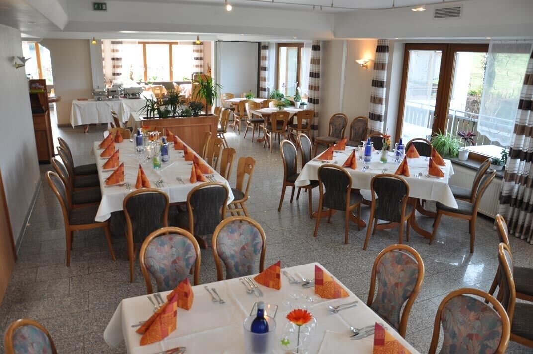 Un espace repas avec plusieurs tables dressées pour les convives, agrémenté de nappes blanches et de serviettes orange.
