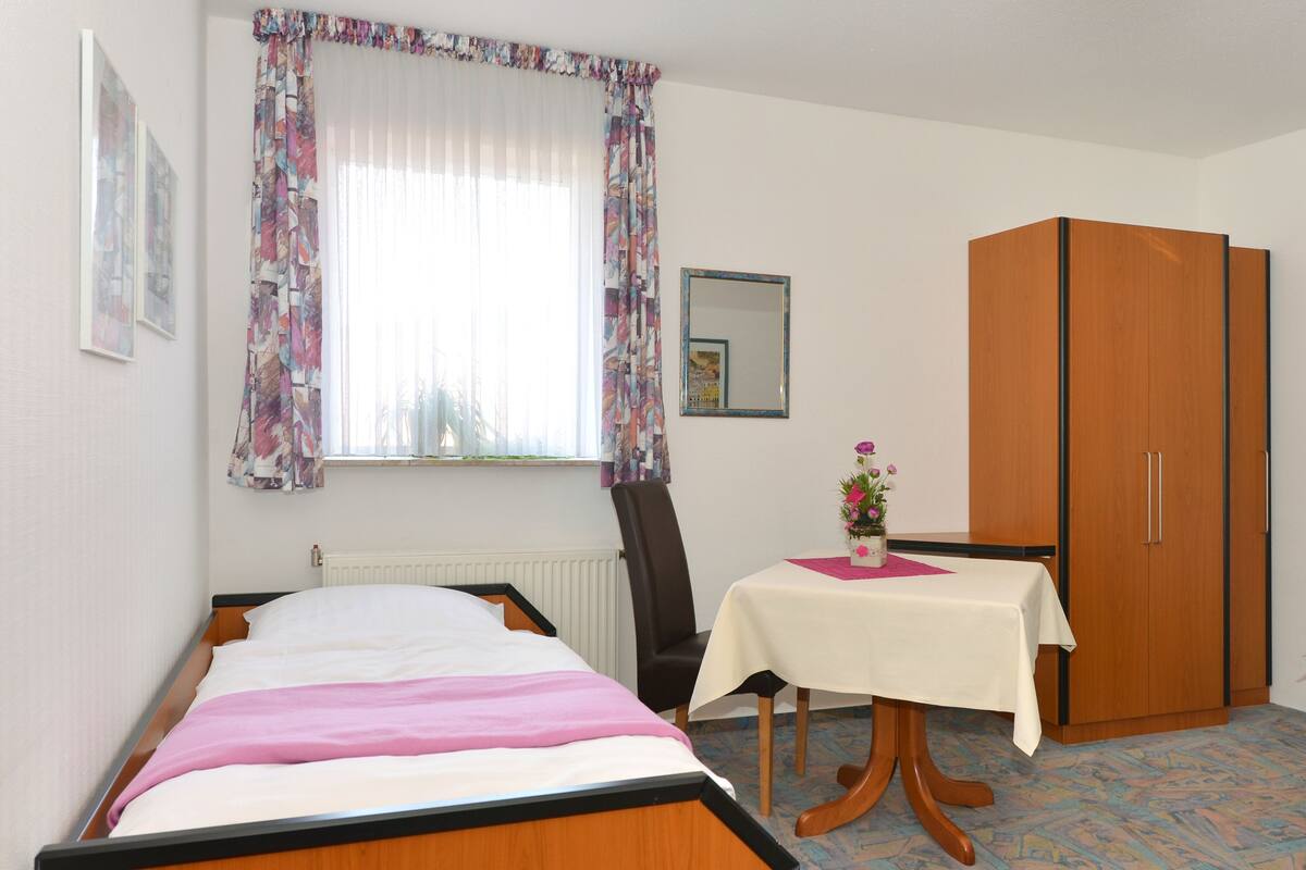 Un dormitorio con cama, armario de madera, mesa con un florero y ventana con cortinas.