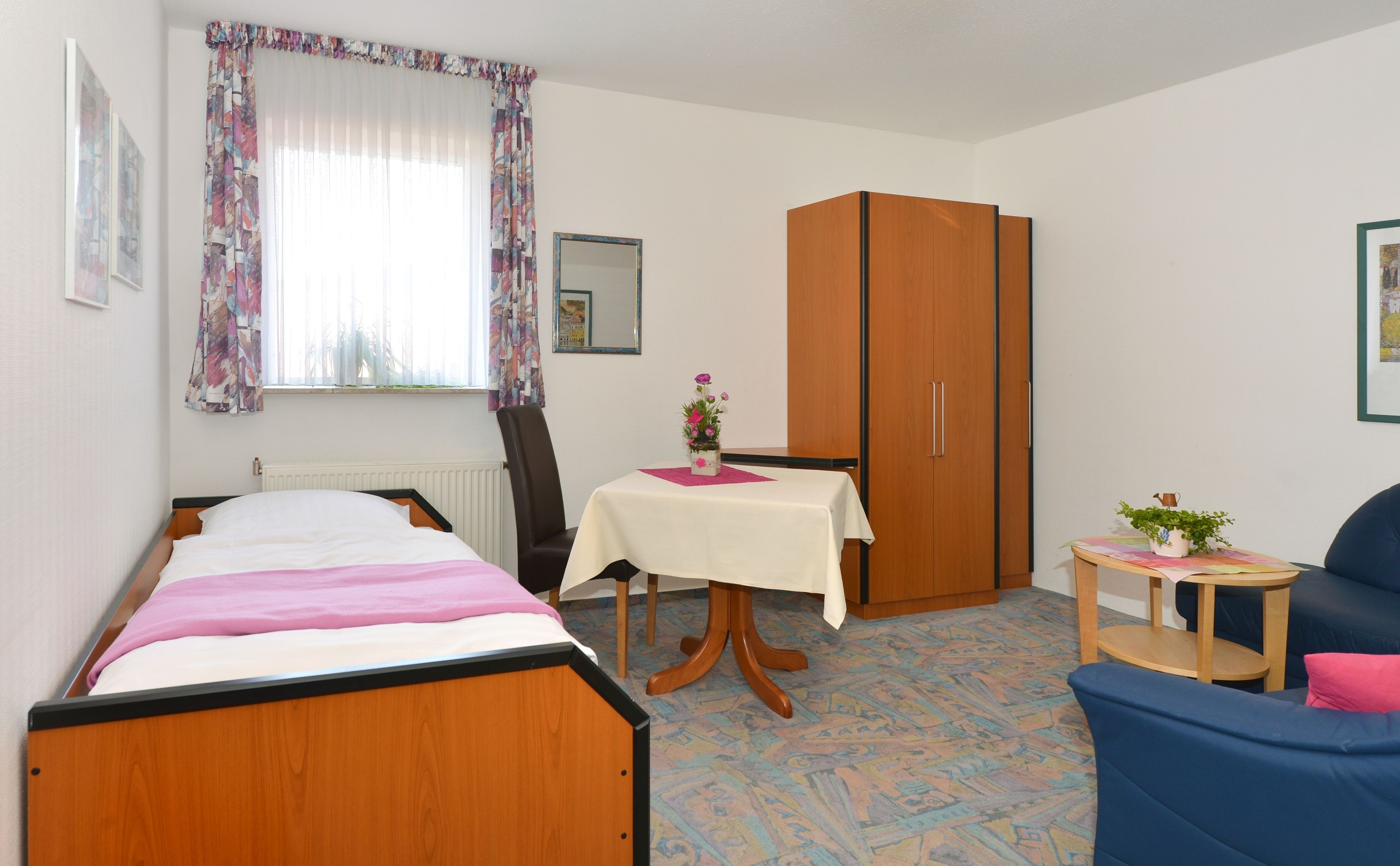 A bedroom with a bed, a chair, a table with a vase, a wardrobe, and a window with curtains.