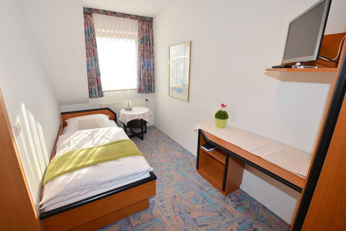 Una habitación pequeña y bien iluminada con cama, escritorio, silla, mesita, radiador, televisor y ventana con cortinas.