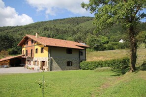 A two-story house with a stone facade and a tiled roof, surrounded by a grassy yard and a forested hillside.