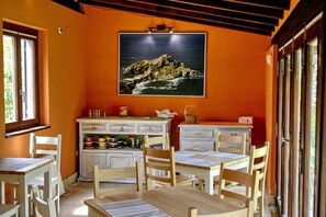 A dining area with wooden tables and chairs, a kitchen counter with dishes, and a large painting of a coastal landscape on the wall.