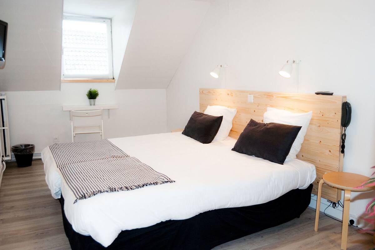 A modern bedroom with a large bed, a bedside table, a small desk, a window, and a telephone.