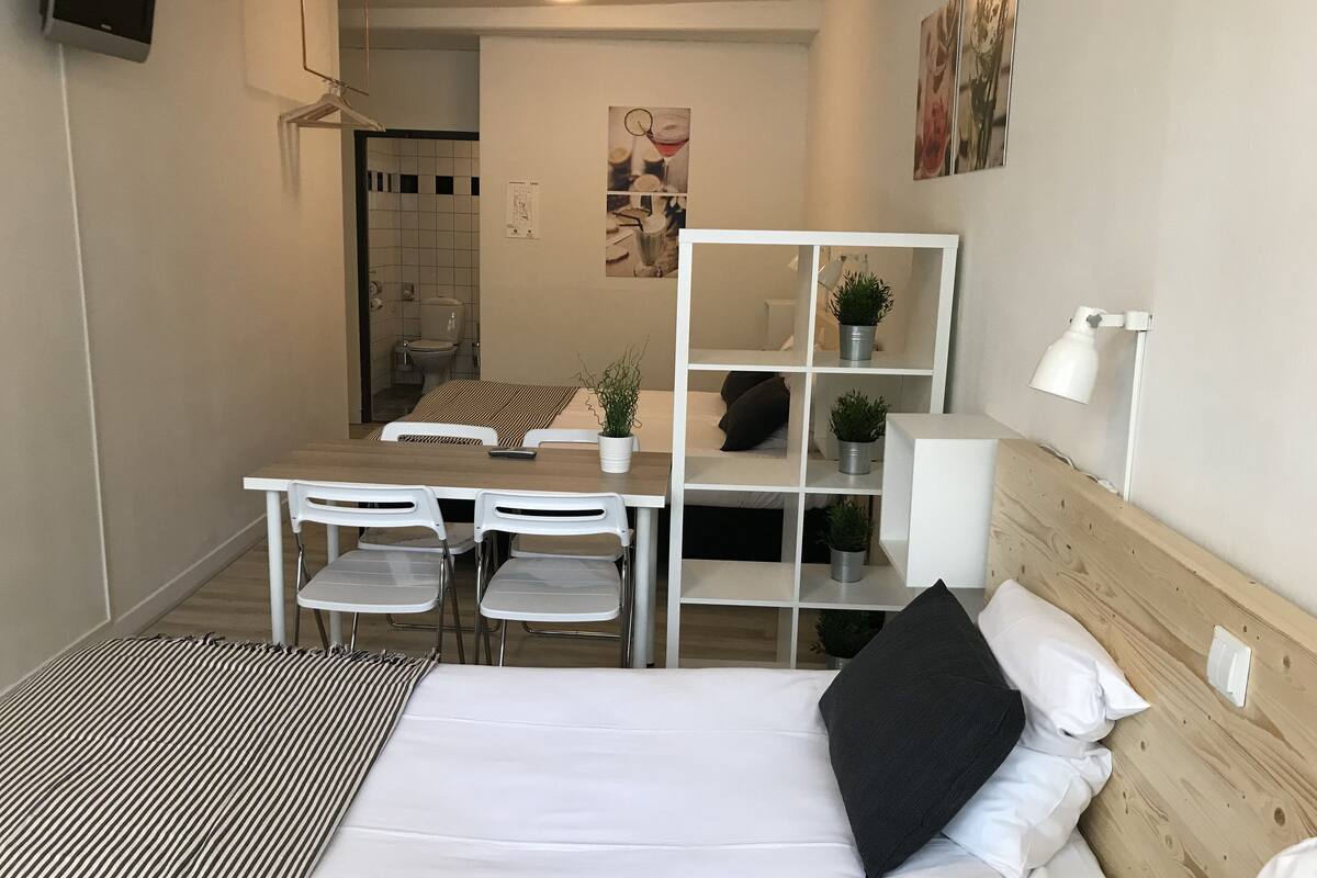 A compact hotel room with a bed, a small table with chairs, a white shelf with plants, and a white lamp.