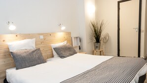 Een net opgemaakt bed met een gestreept dekbed, twee kussens en een houten hoofdbord. Er staat een nachtkastje met een lamp en een plant in een pot.