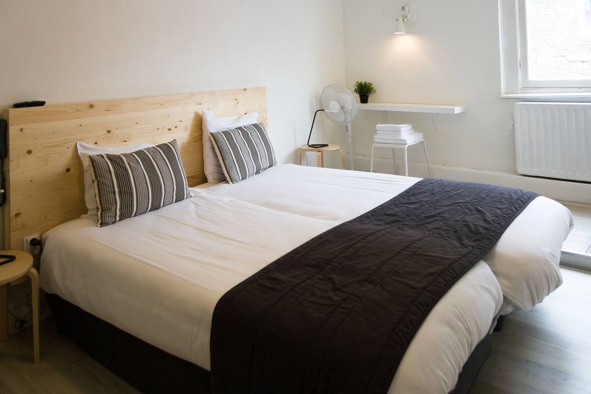 A neatly made bed with a wooden headboard, two pillows, and a black throw blanket. There is a small bedside table with a lamp and a white chair. A white radiator and a small plant are visible on the wall.