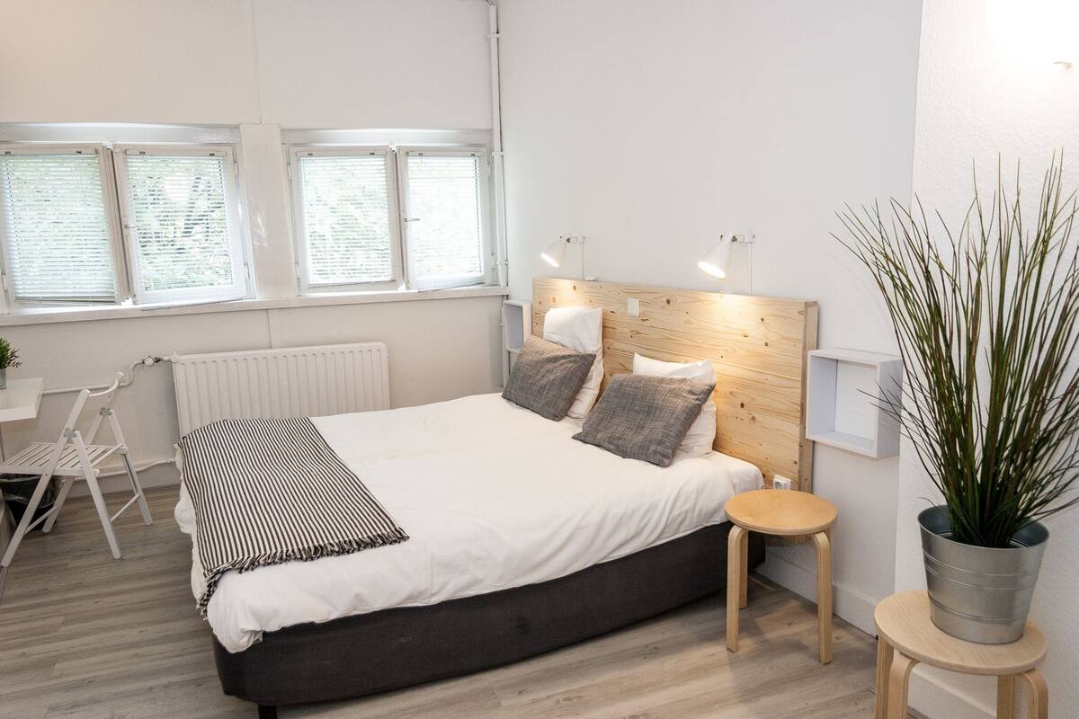 A modern bedroom with a large bed, bedside tables, a radiator, and a window with blinds.