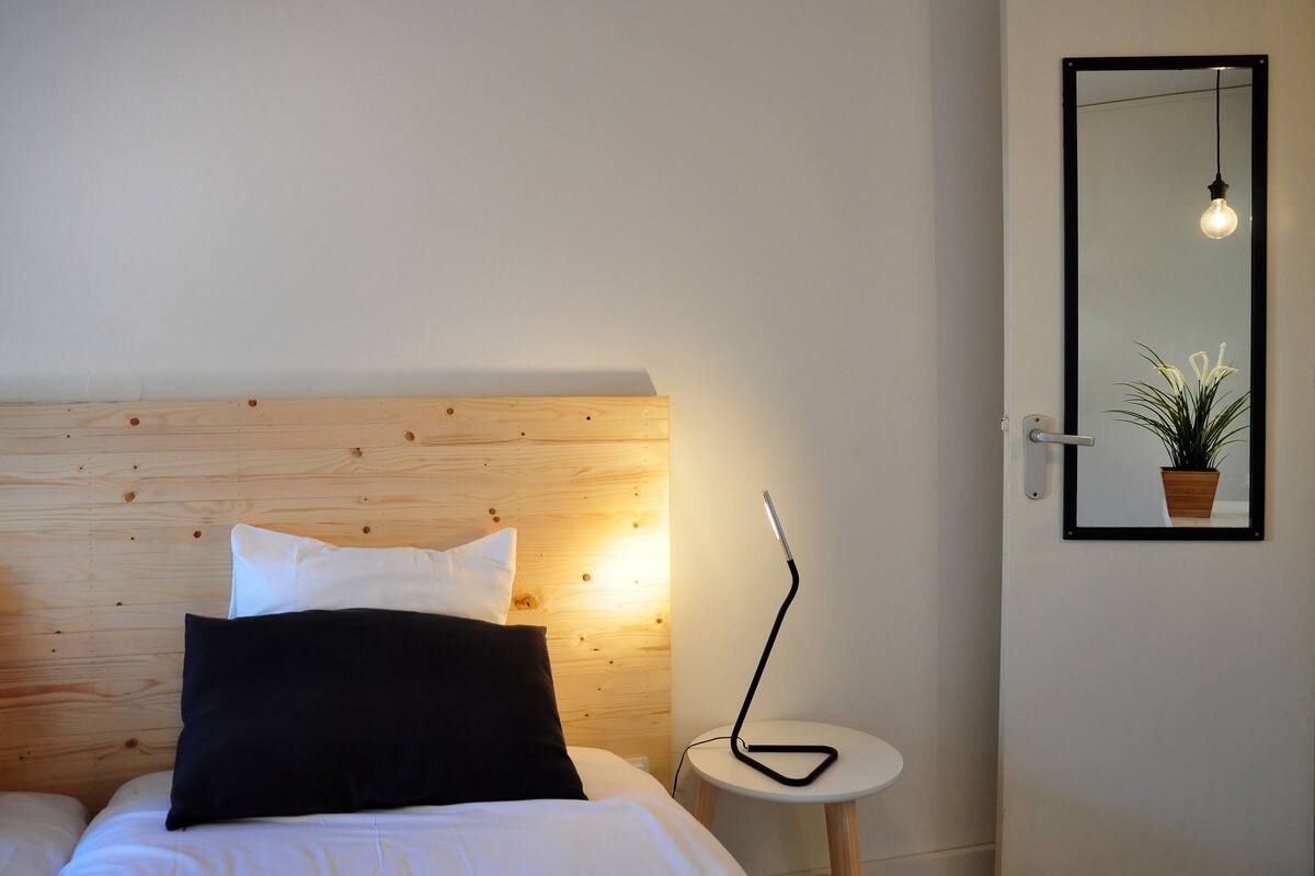 A bedroom with a wooden headboard, a bedside lamp, a round nightstand, and a mirror on the wall.