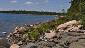 A rocky shoreline with a calm lake, surrounded by dense forest and a clear sky. - Bruce Bay Cottages & Lighthouse (Bruce Mines)