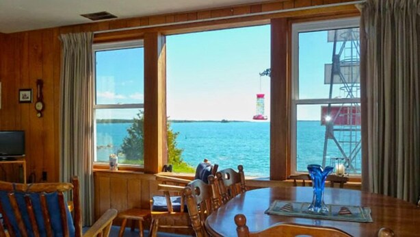 A wooden interior with a large window overlooking a body of water, a television, and a dining area with chairs and a table. - Bruce Bay Cottages & Lighthouse (Bruce Mines)