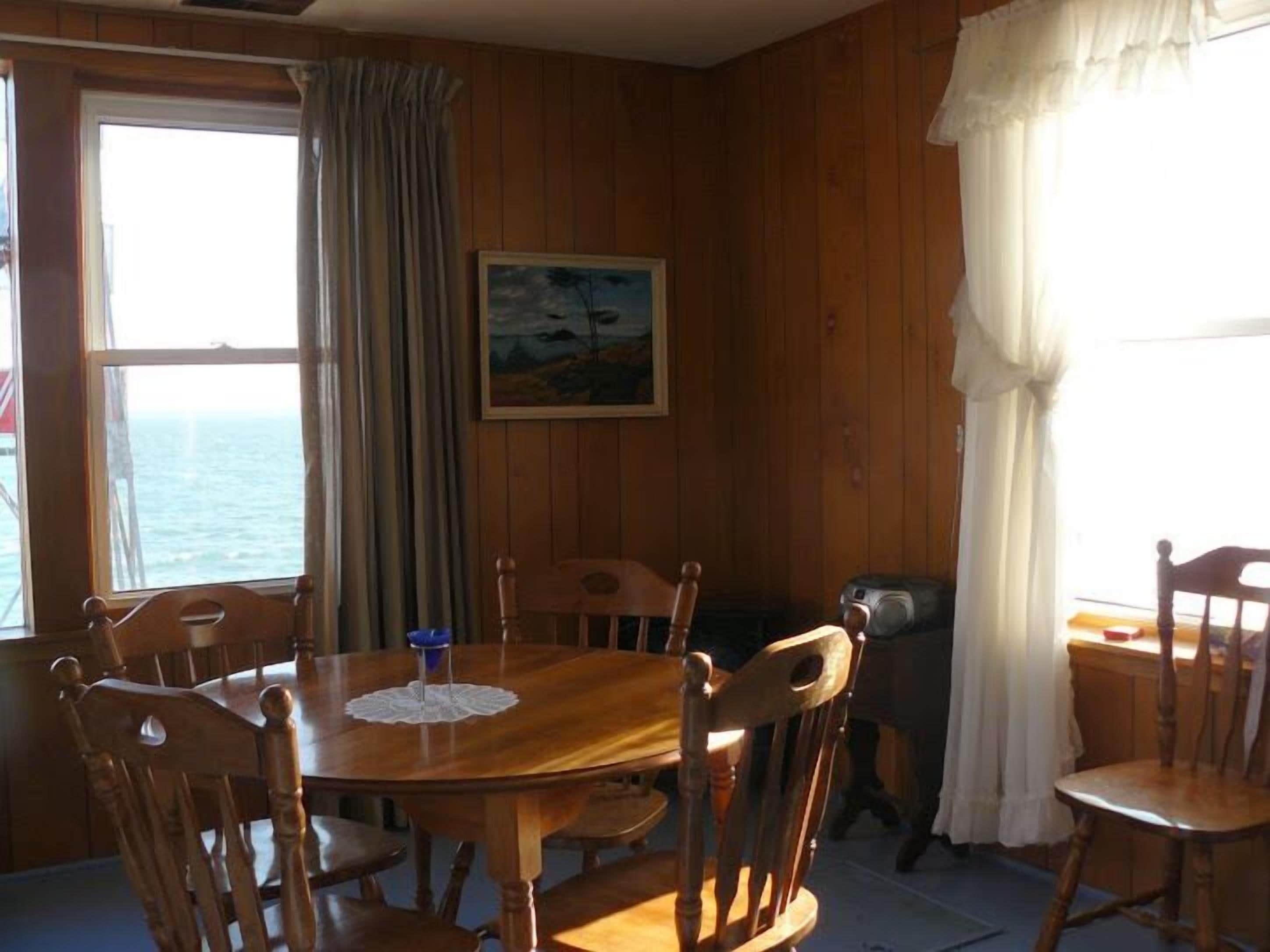Una habitación con paneles de madera, una mesa redonda de madera y sillas, un cuadro en la pared y vista al mar a través de una ventana.