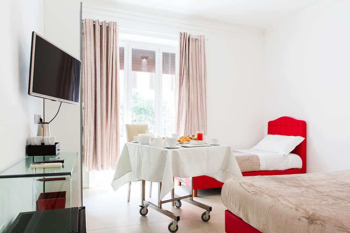 A hotel room with a bed, a desk with a TV, a table set for breakfast, and a window with curtains.