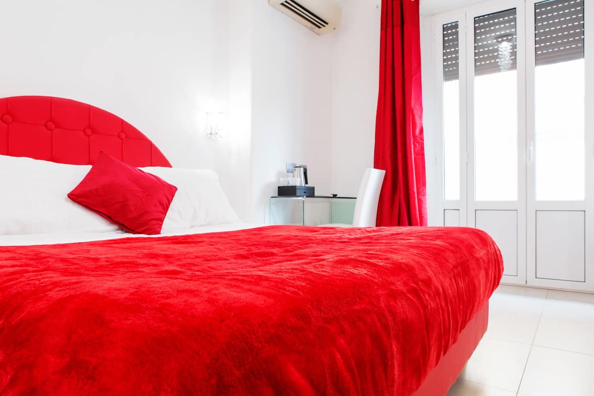 A modern hotel room with a large bed, red bedding, a white headboard, and a view through large windows with red curtains.
