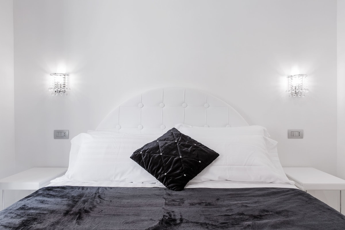 A bed with a white headboard, two bedside lamps, and a black quilted pillow.