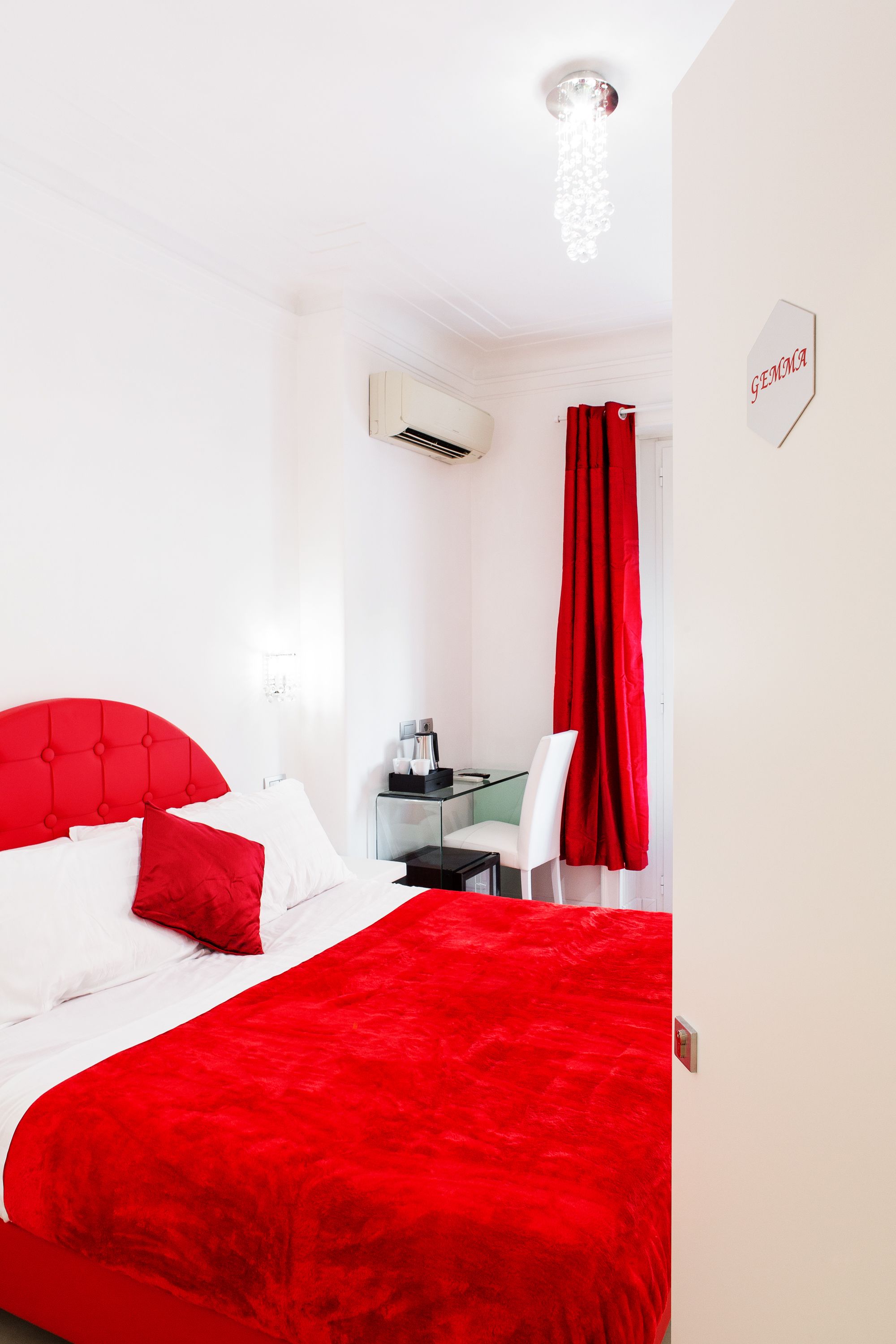 A hotel room with a red bedspread, a white headboard, a desk with a chair, and a red curtain.