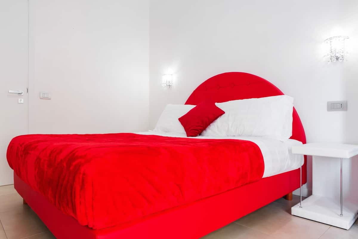 A bed with a red headboard and white bedding, a white bedside table, and a wall-mounted light fixture.
